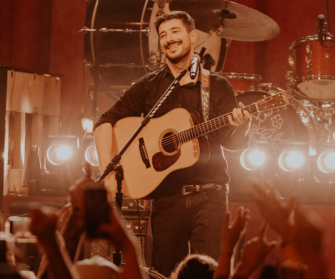 Marcus Mumford, chanteur du groupe Mumford & sons est sur scène et tiens un sa guitar. Il sourit.
